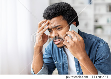 A distressed young adult black man is seen having a serious conversation on a smartphone, expressing concern or confusion A distressed young adult black man is seen having a serious conversation on a smartphone, expressing concern or confusion 114703451