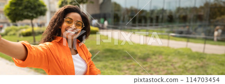 Glad young african american woman in casual, glasses takes selfie on phone on street in city, close up. Photo for social networks, mobility in urban, panorama with copy space Glad young african american woman in casual, glasses takes selfie on phone on street in city, close up. Photo for social networks, mobility in urban, panorama with copy space 114703454