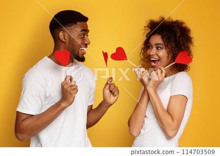Love is around us. Young loving african american couple holding red paper hearts and smiling to each other, orange background Love is around us. Young loving african american couple holding red paper hearts and smiling to each other, orange background 114703509