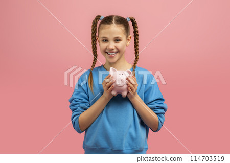 A cheerful young girl with braided hair holding a pink piggy bank and smiling against a pink background 114703519