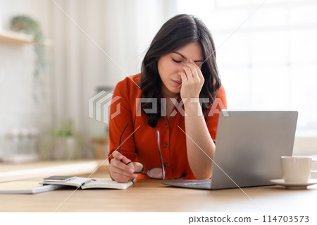 Middle eastern woman appears distressed or exhausted, holding her glasses, with a laptop, notebook, and cup in front of her 114703573