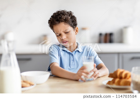 The photograph shows African American boy with a visibly distasteful reaction while holding a glass of milk, suggestive of dislike or dietary discomfort in a kitchen The photograph shows African American boy with a visibly distasteful reaction while holding a glass of milk, suggestive of dislike or dietary discomfort in a kitchen 114703584