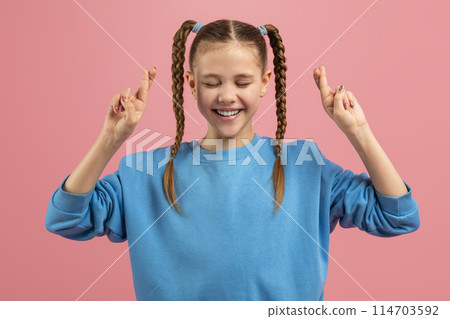 A cheerful young girl with braided pigtails wearing a blue sweater is crossing her fingers hoping for good luck, on a pink background A cheerful young girl with braided pigtails wearing a blue sweater is crossing her fingers hoping for good luck, on a pink background 114703592