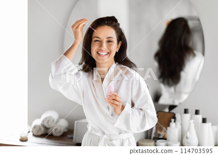 A young woman in a white robe holding a skincare bottle smiles in front of a bathroom mirror, surrounded by toiletry items A young woman in a white robe holding a skincare bottle smiles in front of a bathroom mirror, surrounded by toiletry items 114703595