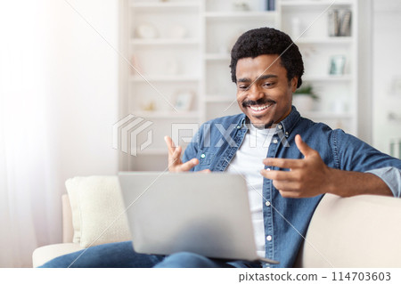 A black african american guy is seen laughing joyfully during a video call at home, interacting with a man on his laptop 114703603