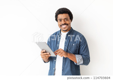 Smiling african american guy uses a tablet at home, showing ease with technology and modern lifestyle Smiling african american guy uses a tablet at home, showing ease with technology and modern lifestyle 114703604