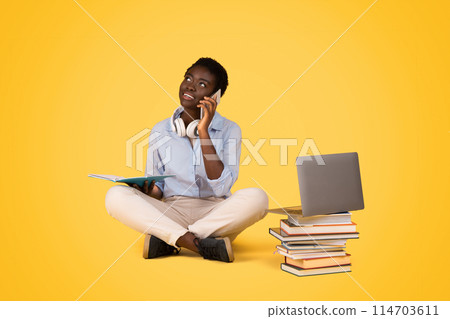 A thoughtful generation z african american sits cross-legged with a book and laptop, contemplating, isolated against a bright yellow backdrop, have phone conversation A thoughtful generation z african american sits cross-legged with a book and laptop, contemplating, isolated against a bright yellow backdrop, have phone conversation 114703611