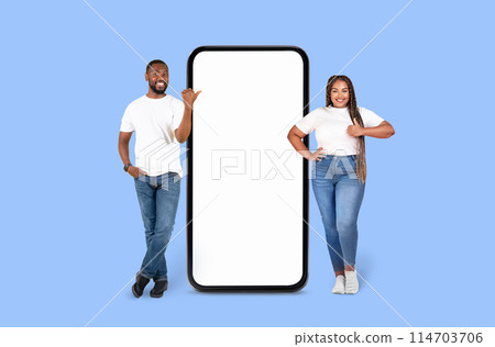 Happy African American pair posing by a large mobile phone display mockup on a blue backdrop, ideal for ads 114703706