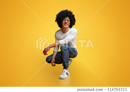 My style. Studio portrait of young black man hunkering down and smiling, orange background 114703721