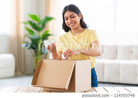 A cheerful muslim woman unpacks white jars cosmetics from a cardboard box, purchasing beauty products online 114703749