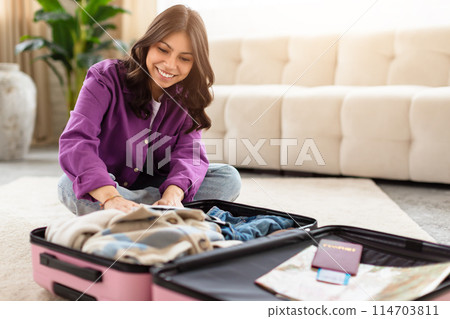 Middle eastern woman in casual clothing carefully arranges items in her pink suitcase at home, implying an upcoming journey or vacation 114703811
