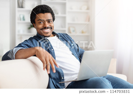 In the comforts of home, an african american man enjoys some leisure time with his laptop, sporting a welcoming smile In the comforts of home, an african american man enjoys some leisure time with his laptop, sporting a welcoming smile 114703831