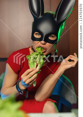 A beautiful girl in a bdsm rabbit mask and a bright red dress eats lettuce leaves promoting a healthy lifestyle and vegetarianism 114703970