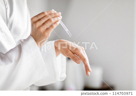 Cropped of woman in robe testing skin care product in bathroom at home, dropping serum on her hand 114703971