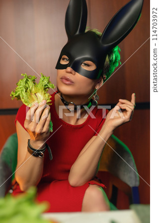 A beautiful girl in a bdsm rabbit mask and a bright red dress eats lettuce leaves promoting a healthy lifestyle and vegetarianism A beautiful girl in a bdsm rabbit mask and a bright red dress eats lettuce leaves promoting a healthy lifestyle and vegetarianism 114703972
