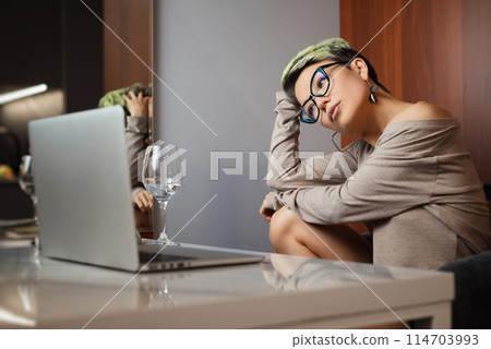 a beautiful girl with short hair and glasses is sitting indoors at a laptop chatting and working online a beautiful girl with short hair and glasses is sitting indoors at a laptop chatting and working online 114703993