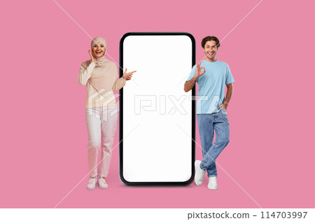 A woman in hijab and man point at blank smartphone screen on a pink background, mockup copy space, mobile app A woman in hijab and man point at blank smartphone screen on a pink background, mockup copy space, mobile app 114703997