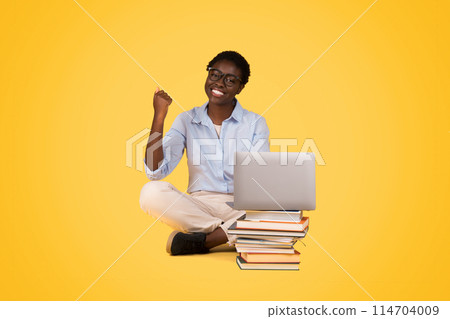 Content African American woman with laptop, seated on a stack of books, showing the joy of learning and the synergy of technology and education, against a yellow background Content African American woman with laptop, seated on a stack of books, showing the joy of learning and the synergy of technology and education, against a yellow background 114704009