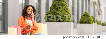 Cheerful young african american woman in casual and glasses with a lot of bags calls by smartphone on city street. Great offer and ad, gossip, good news and communication, copy space, web-banner 114704010