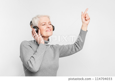 An elderly European woman in a turtleneck smiles while listening to music with headphones, representing joyful s3niorlife 114704033
