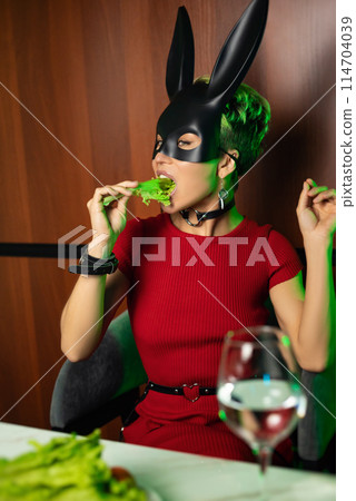 A beautiful girl in a bdsm rabbit mask and a bright red dress eats lettuce leaves promoting a healthy lifestyle and vegetarianism 114704039