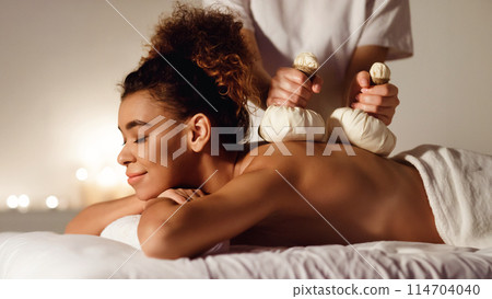 An African American woman enjoys a traditional herbal compress massage at a spa, surrounded by soft lighting and people in the background An African American woman enjoys a traditional herbal compress massage at a spa, surrounded by soft lighting and people in the background 114704040