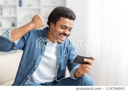 An exuberant african american guy celebrates winning a game or receiving good news on his smartphone at home An exuberant african american guy celebrates winning a game or receiving good news on his smartphone at home 114704041