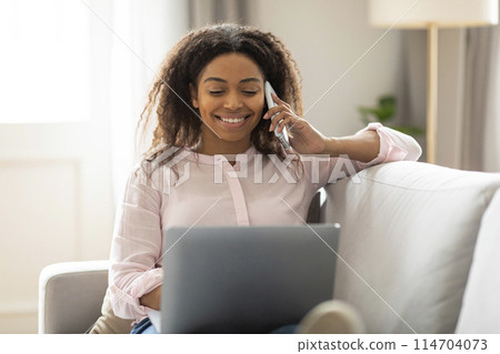 The African American woman is smiling while she talks on the phone and looks at her laptop, showing a home multitasking scenario 114704073