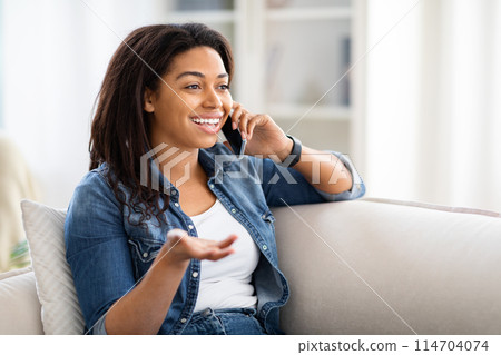African American woman is seated on a couch, engaged in a phone conversation. She holds a cell phone to her ear with focused attention, gesturing occasionally during the call. 114704074