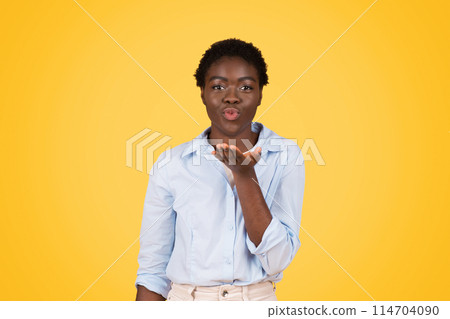 Playful and charismatic black woman sends a flying kiss towards the camera, highlighting her liveliness and Gen Z lifestyle, isolated yellow setting 114704090