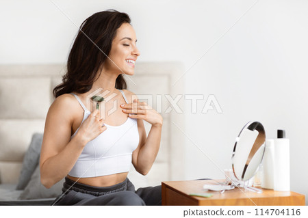 A smiling woman in a white top uses a green jade roller on her skin, sitting in a bedroom with a mirror and products A smiling woman in a white top uses a green jade roller on her skin, sitting in a bedroom with a mirror and products 114704116