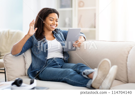 Positive African American woman is seated on a couch, holding a tablet in her hands. She appears focused as she interacts with the device, possibly browsing, reading, or working, have video call 114704140
