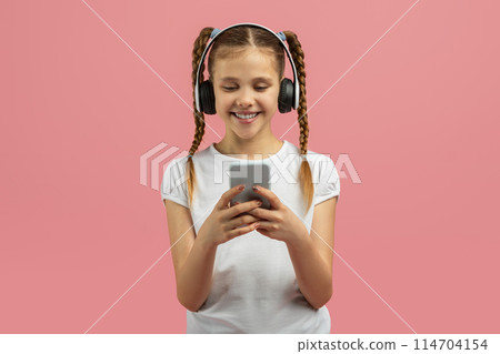 Smiling teen girl with headphones engaged with a smartphone, isolated on pink background, suggesting a connected youngster lifestyle 114704154