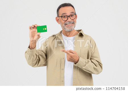 A smart senior man points at a green credit card, possibly suggesting financial options or advantages, isolated on a white background 114704158