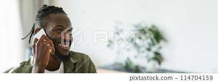 Happy african american man talking on smartphone and smiling, having pleasant phone conversation at home. Mobile communication, technology and gadgets concept, panorama with copy space 114704159