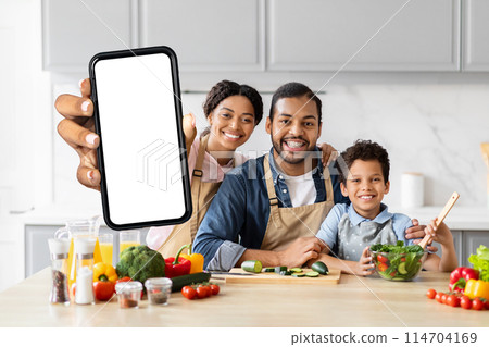 Positive African American family cooking together at kitchen, showing smartphone with white blank screen, recommending food blog Positive African American family cooking together at kitchen, showing smartphone with white blank screen, recommending food blog 114704169