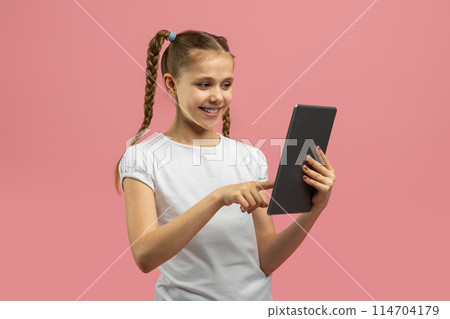 A cheerful young girl with braided hair is interacting with a tablet against a vibrant pink background, looking engaged 114704179