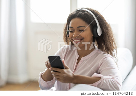 A cheerful young black woman wearing over-ear headphones is focused on her smartphone screen, possibly choosing music or texting A cheerful young black woman wearing over-ear headphones is focused on her smartphone screen, possibly choosing music or texting 114704218