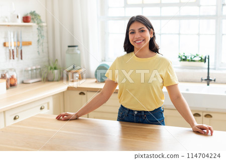 A cheerful arab woman with a bright smile stands confidently in her home kitchen, presenting a warm and inviting atmosphere 114704224