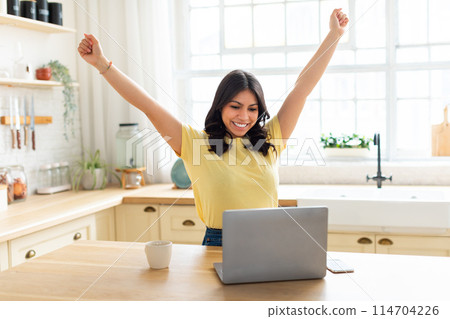 Excited arab woman with arms raised in victory while working on laptop in her cozy kitchen, blending modern comforts with middle eastern aesthetic at home Excited arab woman with arms raised in victory while working on laptop in her cozy kitchen, blending modern comforts with middle eastern aesthetic at home 114704226