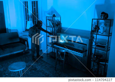 A burglar stands by a piano in an apartment, evaluating items to steal. The blue tint conveys a feeling of isolation at night A burglar stands by a piano in an apartment, evaluating items to steal. The blue tint conveys a feeling of isolation at night 114704227