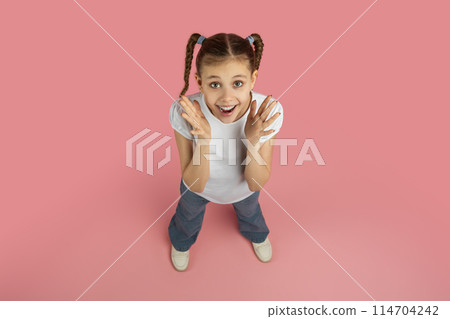 A joyful young girl wearing a white shirt and blue jeans with pigtails smiling and standing on a pink background, expressing positivity and happiness, top view A joyful young girl wearing a white shirt and blue jeans with pigtails smiling and standing on a pink background, expressing positivity and happiness, top view 114704242