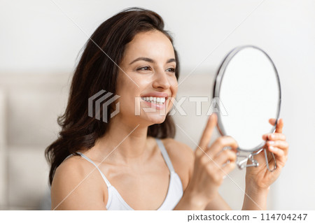 Content millennial brunette pretty lady examines her clear skin, reflecting joy and confidence in a bathroom Content millennial brunette pretty lady examines her clear skin, reflecting joy and confidence in a bathroom 114704247