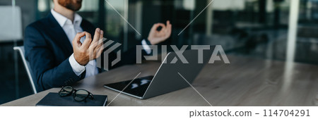 Relaxed businessman meditating at workplace in office, calm man entrepreneur in suit sitting at desk and coping with stress at work, cropped, panorama with copy space 114704291