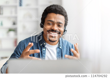 A joyful African American man wearing a headset is engaging enthusiastically with someone or something on his laptop screen in a bright room A joyful African American man wearing a headset is engaging enthusiastically with someone or something on his laptop screen in a bright room 114704292