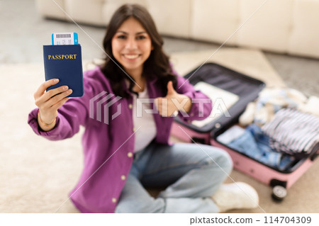 A smiling young middle eastern woman is sitting on the floor with an open suitcase, holding a passport in one hand and giving a thumbs up with the other A smiling young middle eastern woman is sitting on the floor with an open suitcase, holding a passport in one hand and giving a thumbs up with the other 114704309
