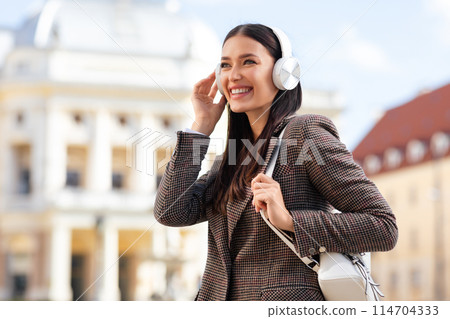 A millennial lady listens to music on headphones, carrying a bag, outdoors with a European building behind her A millennial lady listens to music on headphones, carrying a bag, outdoors with a European building behind her 114704333