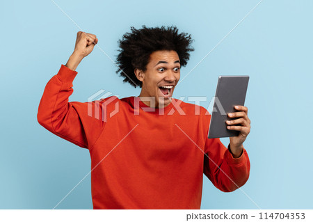 An ecstatic african american guy with a digital tablet celebrating, isolated on blue, ideal for expressing success and achievement 114704353