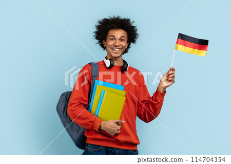 Smiling african american student with bushy hair, posing with the German flag and colorful books, representing international studies or travel, on a blue isolated background 114704354