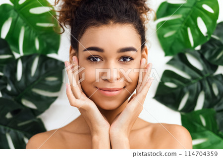 Bright and lively shot of an african american woman at a spa setting with hands on her cheeks and a joyful expression among tropical leaves Bright and lively shot of an african american woman at a spa setting with hands on her cheeks and a joyful expression among tropical leaves 114704359